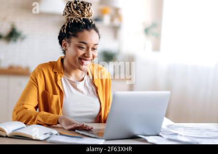 Smart attractive happy mixed race black girl with dreadlocks, in casual clothes, studying remotely, watching online briefing, talking on conference call with teacher or colleagues, smiling Stock Photo