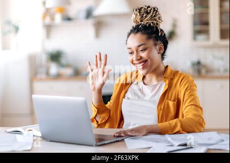 Happy successful beautiful stylish african american girl, student or freelancer, talking by video call, online meeting with colleagues or friends, greeting with hand gesture, smiling friendly Stock Photo