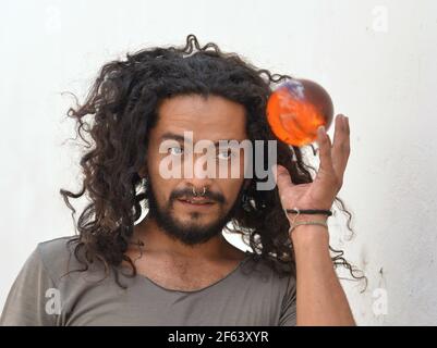 Handsome bearded young Mexican man with long natural curls and septum nose jewelry catches an orange plastic ball with his left hand. Stock Photo