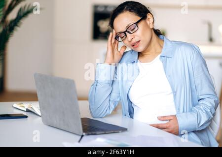 Tired, overworked sad pregnant brunette mature woman wearing glasses, mixed race, freelance, broker or manager, working remotely, took a break, need sleep, sitting at table with eyes closed Stock Photo
