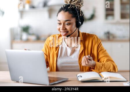 Friendly young attractive African American woman in headset, call center employee, support operator, working at a laptop, conducts online consultation with customer, looking at the screen, smiling Stock Photo