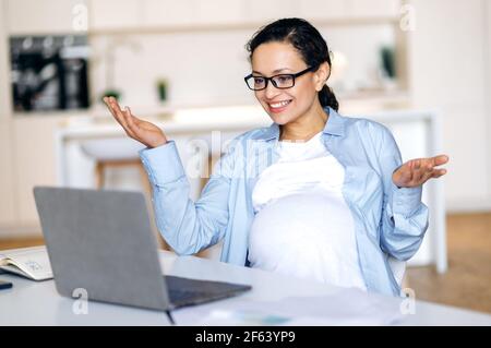 Intelligent successful pregnant mixed race woman, coworker, business lady, manager uses laptop, communicates via video conference, conducts virtual meeting with client or employee, gesture hands,smile Stock Photo