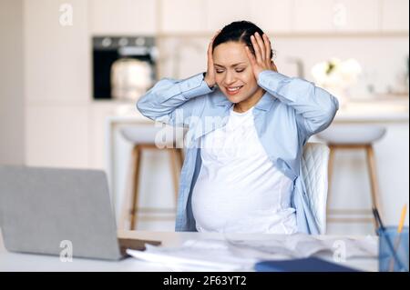 An irritated stressed emaciated mixed race adult pregnant woman, manager, designer or freelancer working remotely, experiencing stress and headache, needing rest, hands holding on her head eyes closed Stock Photo