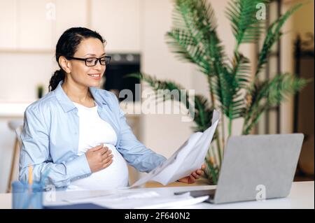 Confident influential pregnant mixed race woman wearing glasses, manager, business lady or lawyer, working at home at table, checking business report, holding documents in hand, smiling Stock Photo