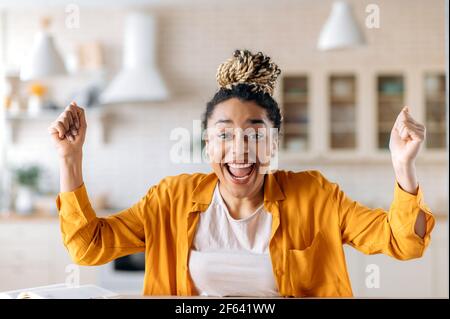 Portrait of joyful attractive confident young African American woman with dreadlocks, in casual clothes, working remotely, emotional face expression, gesturing with hands, rejoices of good job or news Stock Photo