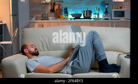 Man falling asleep in fron of TV while working on the laptop. Cozy man lying down on sofa while typing on laptop computer for social media project. Exhausted man working Stock Photo