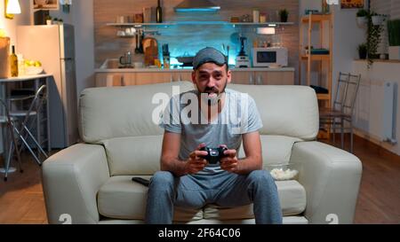 Upset pro gamer sitting in front of television losing soccer video games holding wireless controller. Frustrated man with sleep mask lying on sofa late at night in kitchen Stock Photo