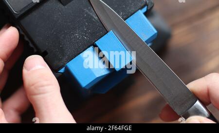 Sharpening a knife on an electric sharpener at home. The man's hand drives the knife blade between the blue sharpeners, dust flies on the machine. Stock Photo