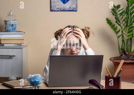 Teenage girl clasping her head in her hands and stares intently at the laptop screen. excited serious teenage girl with glasses is engaged online on a Stock Photo