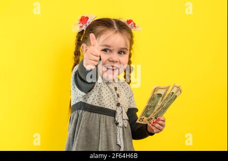 The child shows a thumb up. Children and financial responsibility, little girl on a yellow background with dollars in her hands, financial literacy in Stock Photo