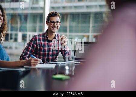 Smiling young man sitting in boardroom meeting. Male professional in meeting with colleagues and smiling, Stock Photo