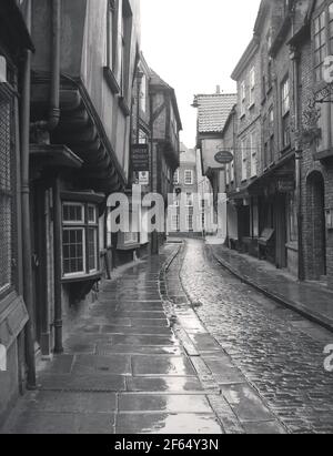 1956, historical, on a wet, damp British day, a view along an empty Shambles, a narrow, cobbled medieval street in York, England, UK, with overhanding timber-frame buildings, some dating as far back to the fourteenth century. Once known as 'The Great Flesh Shambles', from the meat that the butchers used to display in their shop windows, the ancient narrow street is perhaps Europe's best-preserved medieval street. The name 'Shambles' is now commonly used to collectively refer to the surounding maze of narrow twisting lanes and alleys as well. A sign for the York Weavers can be seen. Stock Photo