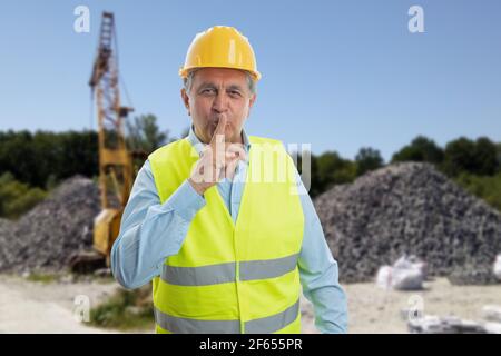 Old mature constructor man wearing yellow protective hardhat and fluorescent vest making shush silence gesture with index finger over lips on construc Stock Photo