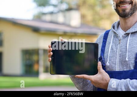 Cropped shot of bearded male builder smiling at camera, holding digital tablet pc with blank screen while working at cottage construction site Stock Photo