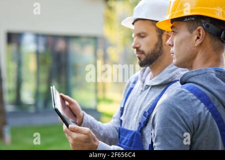 Close up of two young male builders using digital tablet pc, discussing ideas while working at cottage construction site Stock Photo