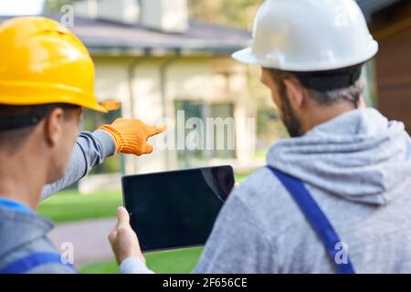 Use your vision. Close up of two young male builders using digital tablet pc, discussing ideas while working at cottage construction site Stock Photo