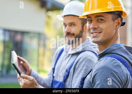 We will get it done. Portrait of two young male builders smiling at camera, using digital tablet pc while working at cottage construction site Stock Photo