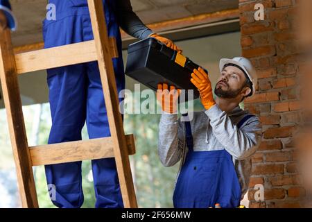 Attractive young male builder in overalls and hard hat giving toolbox to his colleague on ladder while working at cottage construction site Stock Photo