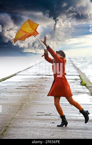 A girl in orange coat with umbrella in stormy weather on the pier. Cloudy sky. Stock Photo