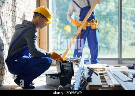 Build it. Young male engineer taking instrument from toolbox while working at cottage construction site with blurred colleague in the background on a Stock Photo