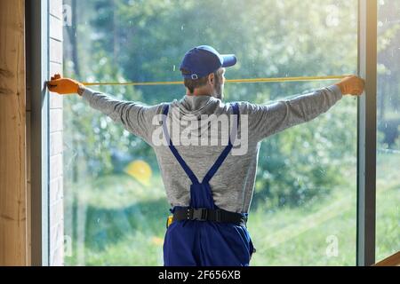 Rear view of young workman wearing blue overalls using measuring tape while working on cottage construction site Stock Photo