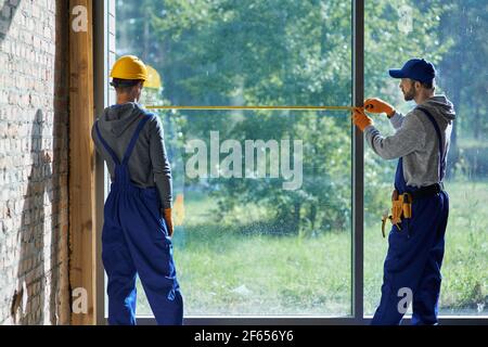 Making new home. Rear view of two young male builders wearing blue overalls using measuring tape while working on cottage construction site Stock Photo