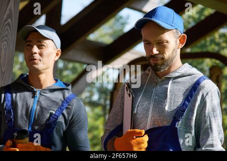 Portrait of two confident young repairmen in uniform standing outdoors with papers while working on construction project Stock Photo