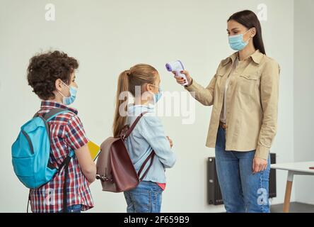 Young female teacher wearing protective mask measuring temperature, screening kids with digital thermometer while students coming to school Stock Photo