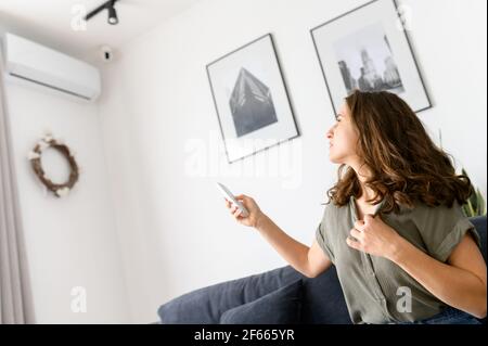 Female using the remote control adjusts the temperature of the air conditioner, young woman suffers from heat at home, switches conditioner on. Technology that improves everyday life Stock Photo