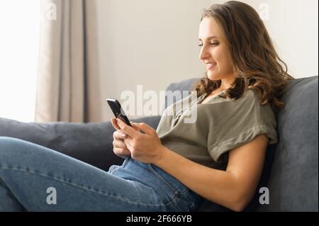 Satisfied young woman with smartphone on the couch rests at home, leaned back in relaxed pose, scrolling news, chatting in social media with friends, messaging online, chatting in date app. Side view Stock Photo