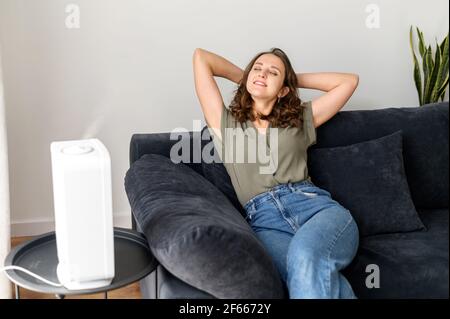 Smiling and serene young woman rests on the comfortable couch under stream of fresh and cleaned air from humidifier in cozy living room, housewife leaned back with eyeclosed and daydreaming Stock Photo