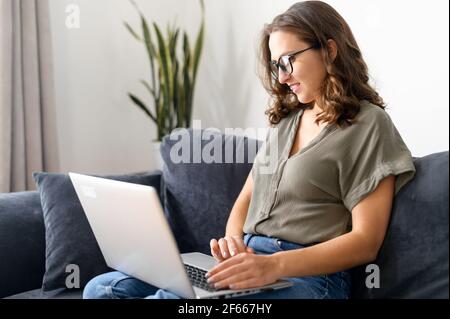 Side view at smiling young woman with a laptop on the couch at home, female entrepreneur using computer for work on the distance, female student learning online Stock Photo