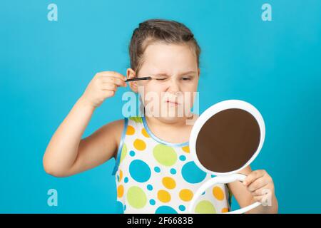 close-up portrait of girl in white dress applying green shiny eye shadow and holding a white plastic mirror in her hand, isolated on a blue background Stock Photo