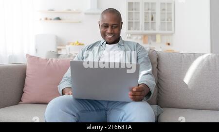 Happy middle-aged black man using laptop while resting on sofa in living room, panorama Stock Photo