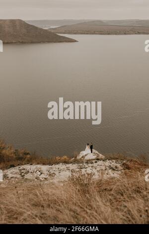 a wedding couple in love, groom and the bride in dress veil stand on Big Stone on cliff and kiss. panoramic view big river lake sea in distance silhou Stock Photo