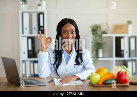 Portrait of black female dietitian showing okay gesture at table with laptop and fresh products in clinic Stock Photo