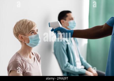 African american male nurse in protective gloves measures temperature with thermometer in adult woman in mask in clinic Stock Photo