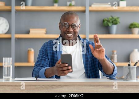 Cheerful mature african american man using smartphone and making greeting gesture, looking and smiling to camera Stock Photo