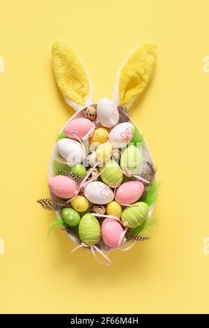 Easter painted eggs in plate as egg and bunny ears on yellow. Vertical Happy Easter greeting card. View from above. Stock Photo