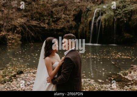 skinny bride in white wedding dress and long veil hugs groom in a suit. small pond lake river. summer-autumn sunset fallen orange leaves mist Stock Photo