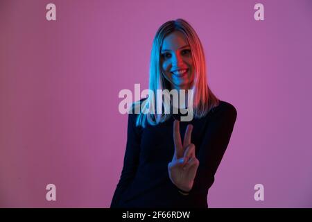 young blonde woman illuminated by neon lights makes the victory sign with her hand Stock Photo