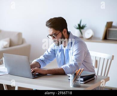 Arab Freelancer Guy Sitting At Desk At Home And Typing On Laptop Stock Photo