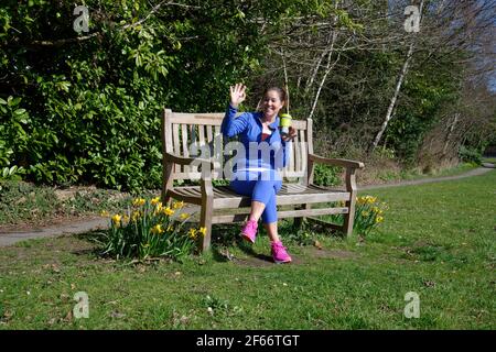 A middle aged woman sitting on a park bench after taking exercise. Cup of coffee in hand and waving to a friend. Stock Photo