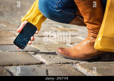 Woman picking up damaged smartphone with cracked touch screen on the street. Broken mobile phone. Stock Photo