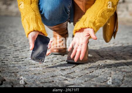 Woman feeling frustration from broken smartphone. Cracked touch screen on mobile phone. Woman picking up damaged smart phone on the street Stock Photo