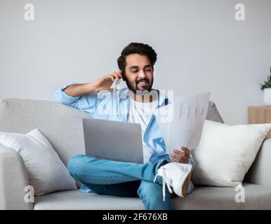 Eastern Freelancer Man With Cellphone, Papers And Laptop Working Remotely At Home Stock Photo