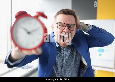 Businessman in glasses holding red alarm clock in his hands and shouting Stock Photo