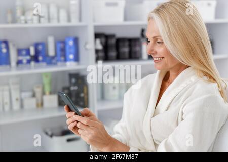 Senior Caucasian female smiling in white bathrobe, holding mobile phone in beautician salon Stock Photo