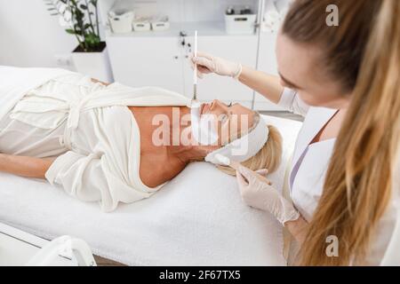 Face peeling mask, spa beauty treatment, skincare. Senior woman getting facial care by beautician at spa salon Stock Photo