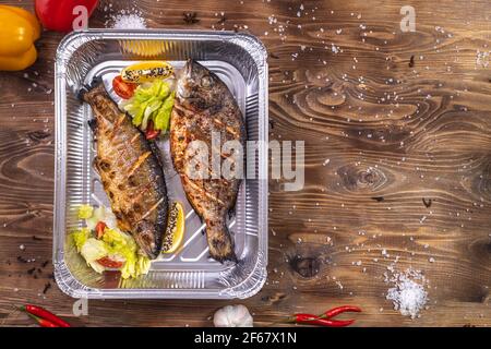 Grilled fish with vegetables, bell pepper, coarse salt, laid in an aluminum container on a brown wooden background. Restaurant food delivery. Stock Photo
